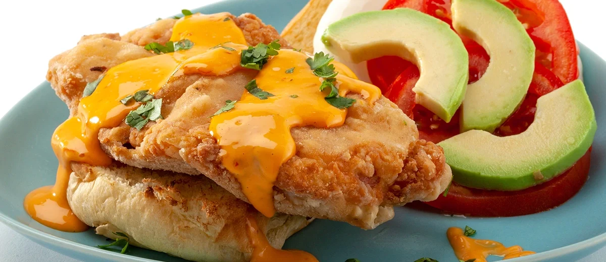 An open face fried chicken sandwich with drizzled orange color sauce, tomato, avocado slices