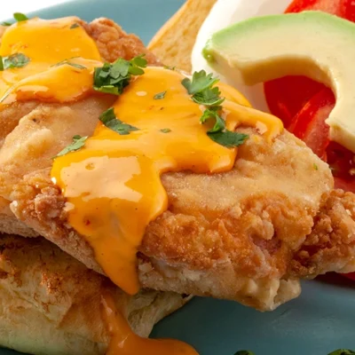 An open face fried chicken sandwich with drizzled orange color sauce, tomato, avocado slices