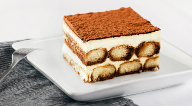 Slice of tiramisu on a white plate with fork and blue gray striped napkin