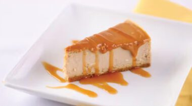 one slice of cheesecake with caramel drizzled over top, on white plate with yellow napkin
