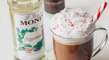 bottle of monin peppermint syrup next to glass cup of hot cocoa with whip cream and peppermint sprinkled on top
