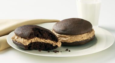 half of eaten peanut butter whoopie pie next to whole peanut butter whoopie pie on white glass plate with glass of milk and brown cloth napkin