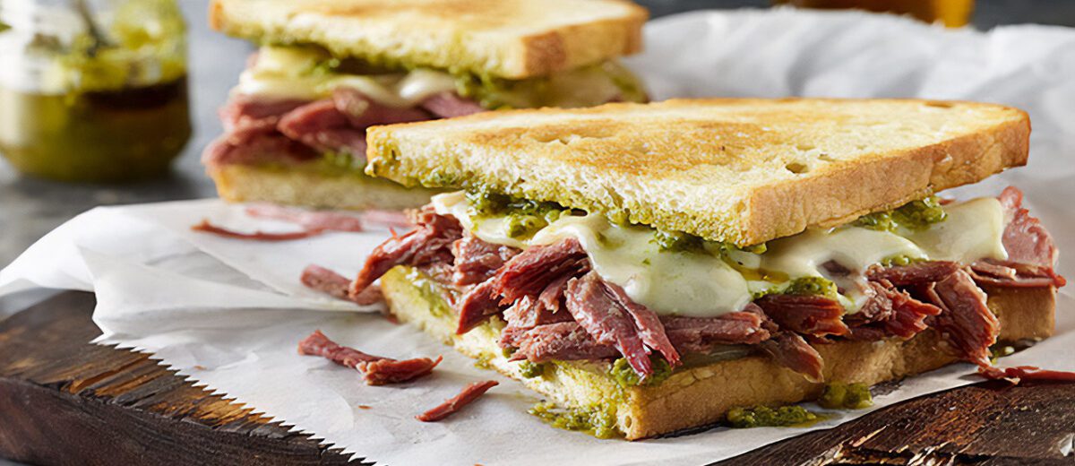 Sandwich, toasted white bread with corned beef pieces, melted cheese and visible pesto