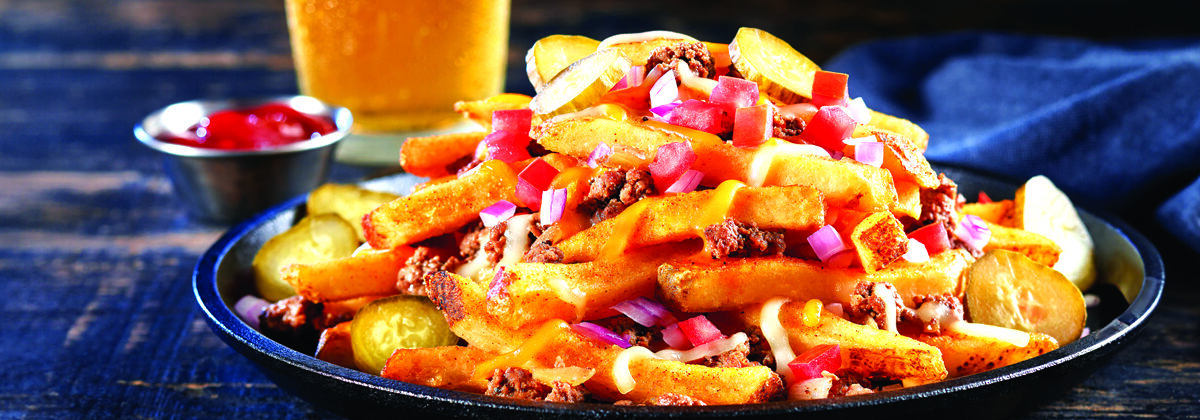 black plate loaded with french fries, hamburger, onions, tomatoes, shredded cheese, peppers ready to eat. ketchu and class of cold beer in background.