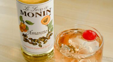 bottle of amaretto syrup next to glass of pink drink with ice and cherry on tabletop