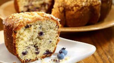 slice of blueberry coffee cake on white plate with two blueberries. Whole cake in background