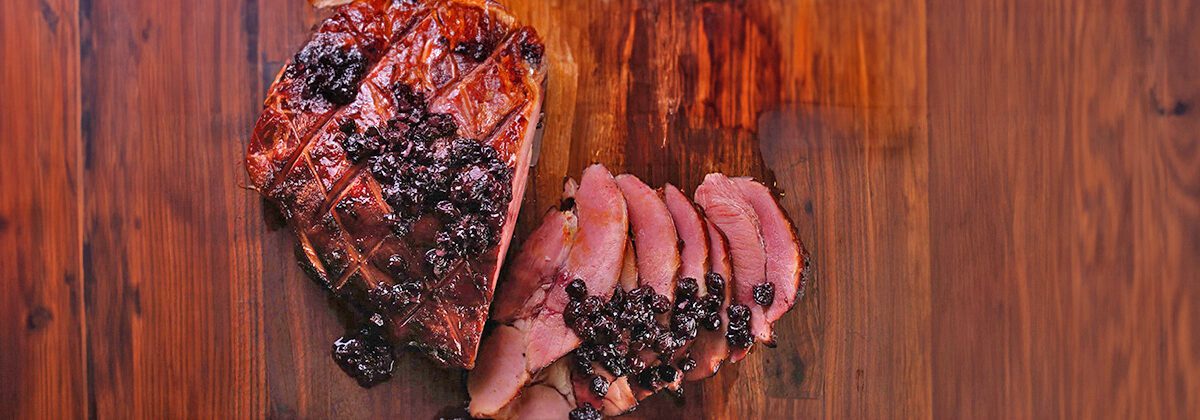 A dark wood cutting board with a big ham, half of which is sliced, and is all covered in a blueberry maple glaze