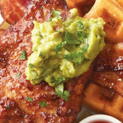 Cooked chicken breast on plate of waffles and bacon with guacamole on top