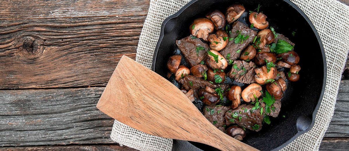 steak tips in a cast iron pan with mushrooms and parsley on a wood background with a wood spoon
