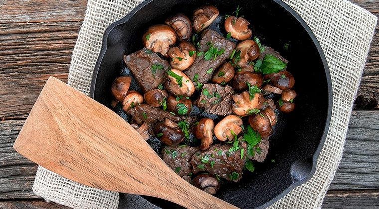 steak tips in a cast iron pan with mushrooms and parsley on a wood background with a wood spoon