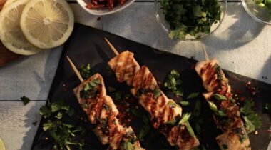 three skewers of cubed salmon sit a on a slate, surrounded by bowls of ingredients and lemon slices, on a tan tablecloth