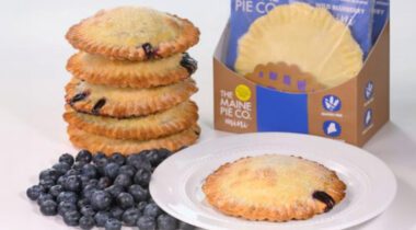 small blueberry pie on white plate beside large pile of blueberries and stack of 5 small blueberry pies and packaged pies.