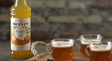 Bottle of Monin Pumpkin Pie Syrup next to 3 glass drinks