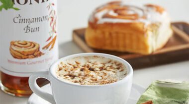 White coffee cup with coffee drink and foamy top and cinnamon sprinkled over top, cinnamon bun in background and Cinnamon Bun Syrup.