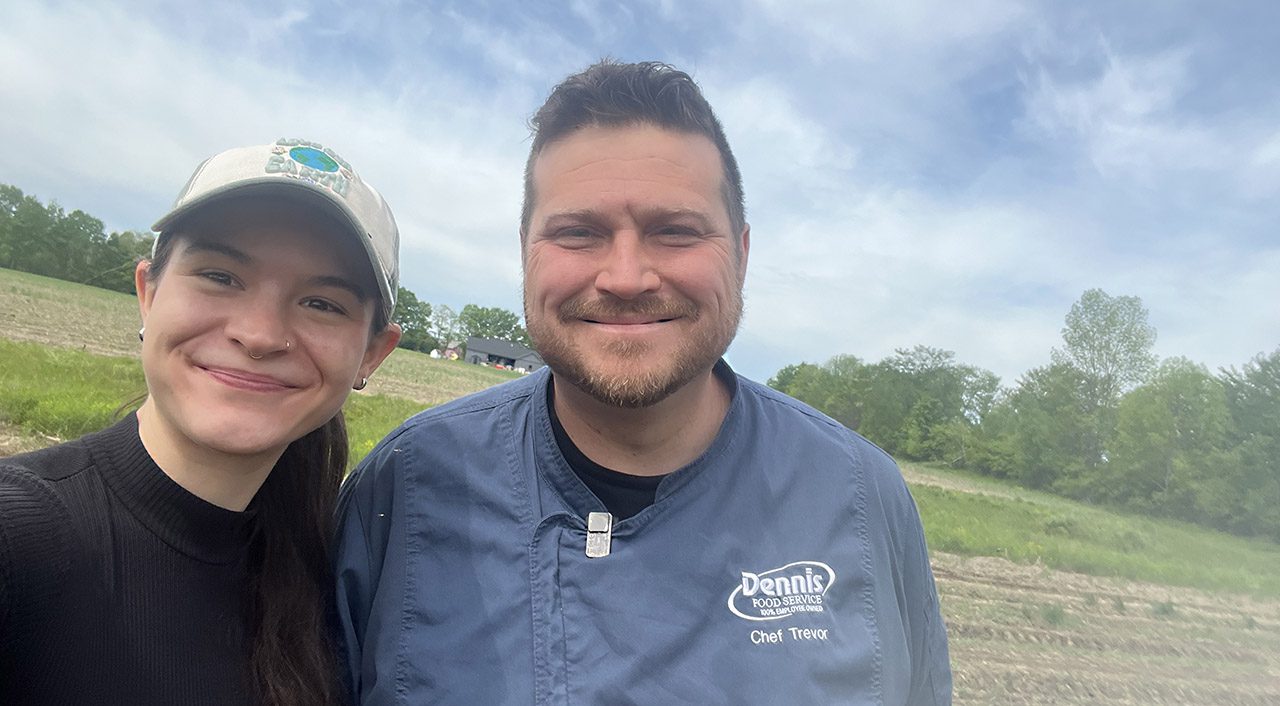 a woman in a black shirt and ball cap and a man in a blue chef jacket take a selfie in a farm field