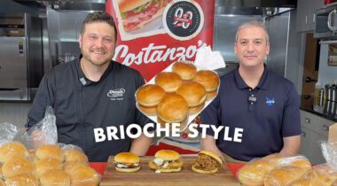 2 smiling people at a table of buns rolls with graphic text says brioche style with signs and marketing for Costanzo's bakery