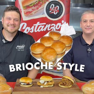2 smiling people at a table of buns rolls with graphic text says brioche style with signs and marketing for Costanzo's bakery