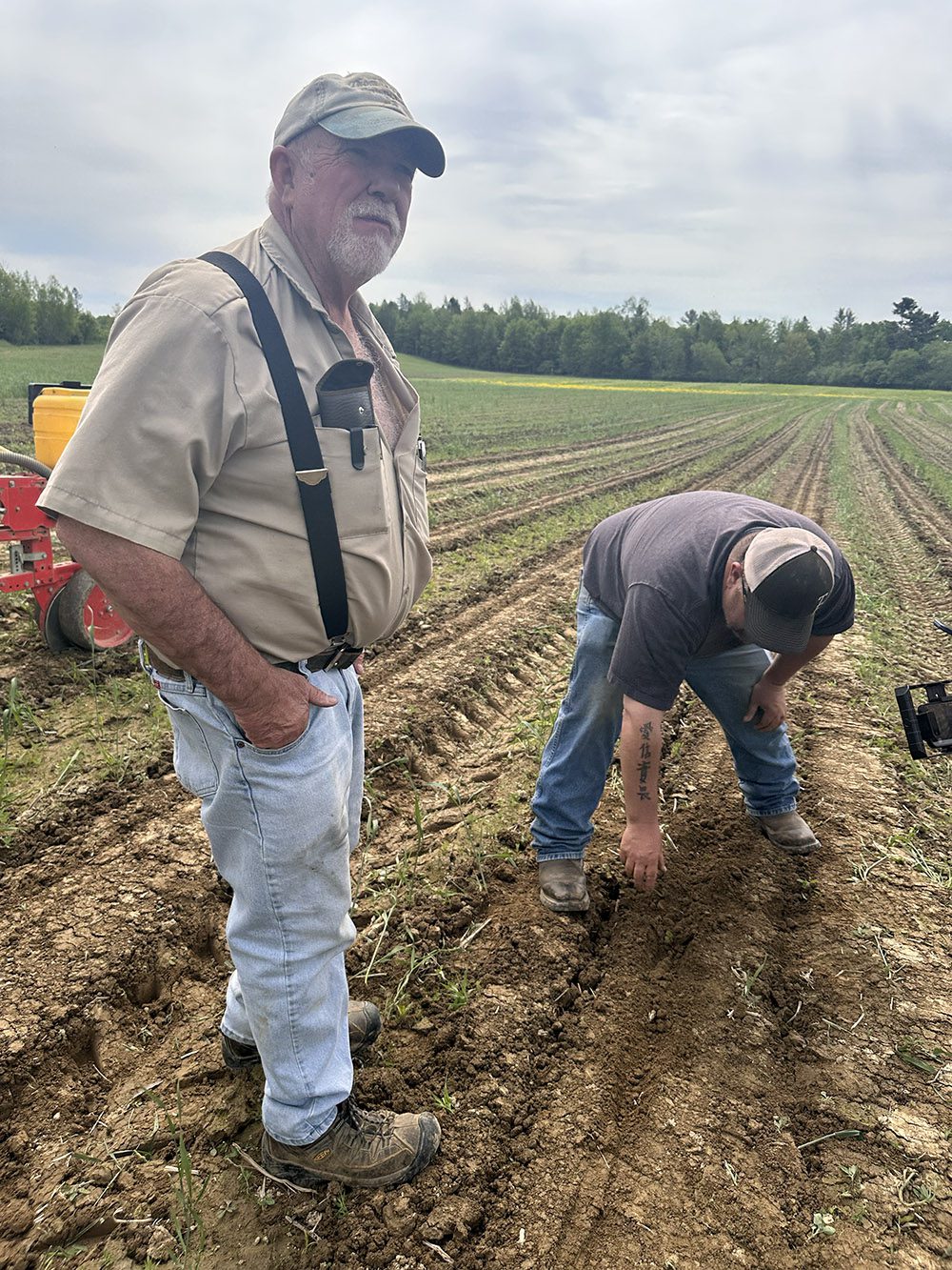 two men in a farm field one is standing, the other stooping to touch soil