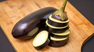 Two fresh, purple Eggplants on wooden cutting board. One sliced and stacked, one whole.