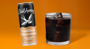 glass of black ice coffee in a glass next to a can with a black and tan label and a white bird on it, orange background