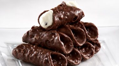 Clear plate of eight chocolate mini cannoli shells. One shell on top filled with cream filling.