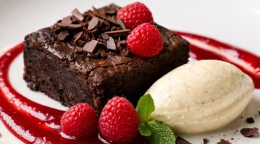 Decadent brownie with raspberry coulis, fresh raspberries, dollop of white ice cream and chocolate shavings on white plate.