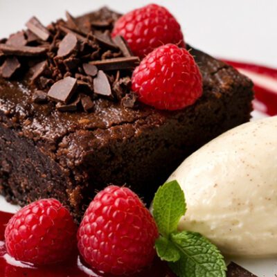 Decadent brownie with raspberry coulis, fresh raspberries, dollop of white ice cream and chocolate shavings on white plate.