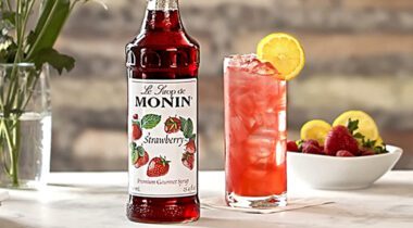 Bottle of Monin Strawberry syrup drink flavor next to tall glass of pink drink with ice and lemon