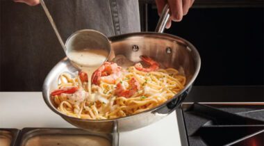 pan of seafood and noodles with ladle drizzling white cream sauce over