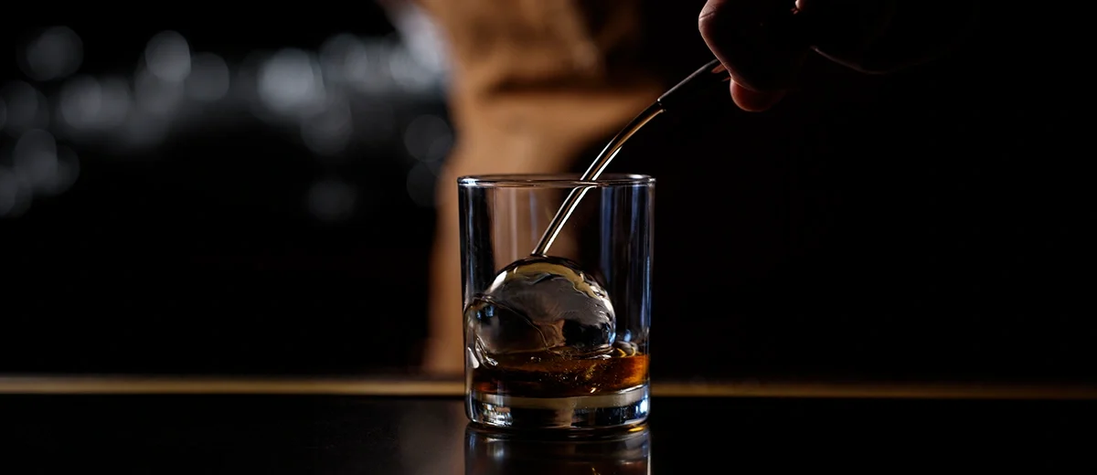 glass with round ice cube and liquor being poured in
