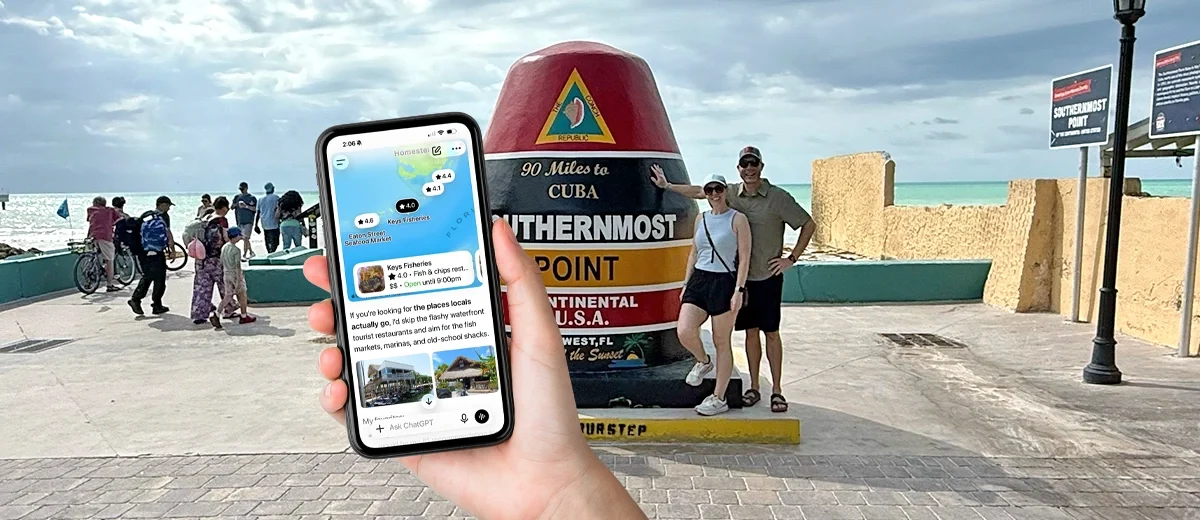 a hand holds a phone out in front of a couple stands at the key west bouy marking the southern most point of the usa